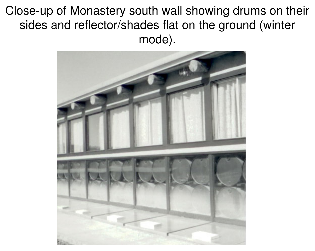 Close-up photograph of the monastery south wall showing rows of steel drums lying on their sides with reflector/shades laid flat on the ground in winter mode