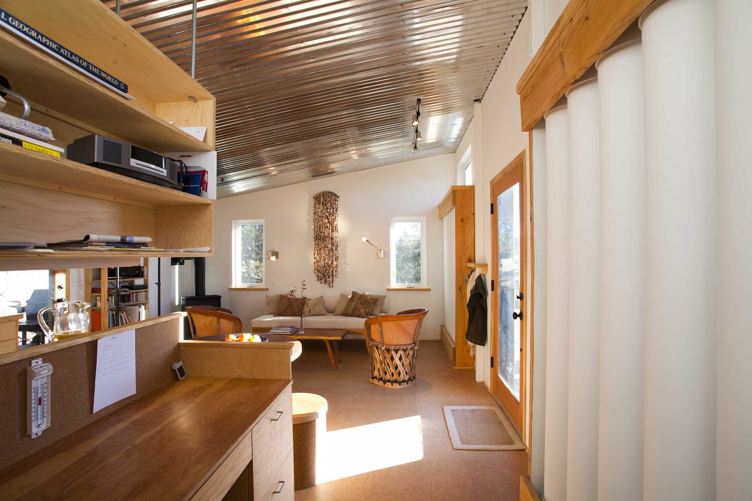 Interior of the Sol-Mod house photographed by Marc Romanelli: a narrow daylit room with a corrugated aluminum ceiling, vertical white water-storage columns along the right wall beside a glazed wood door, built-in birch desk and shelving on the left with a cork bulletin board and thermometer, cork flooring, leather equipal chairs and a sofa