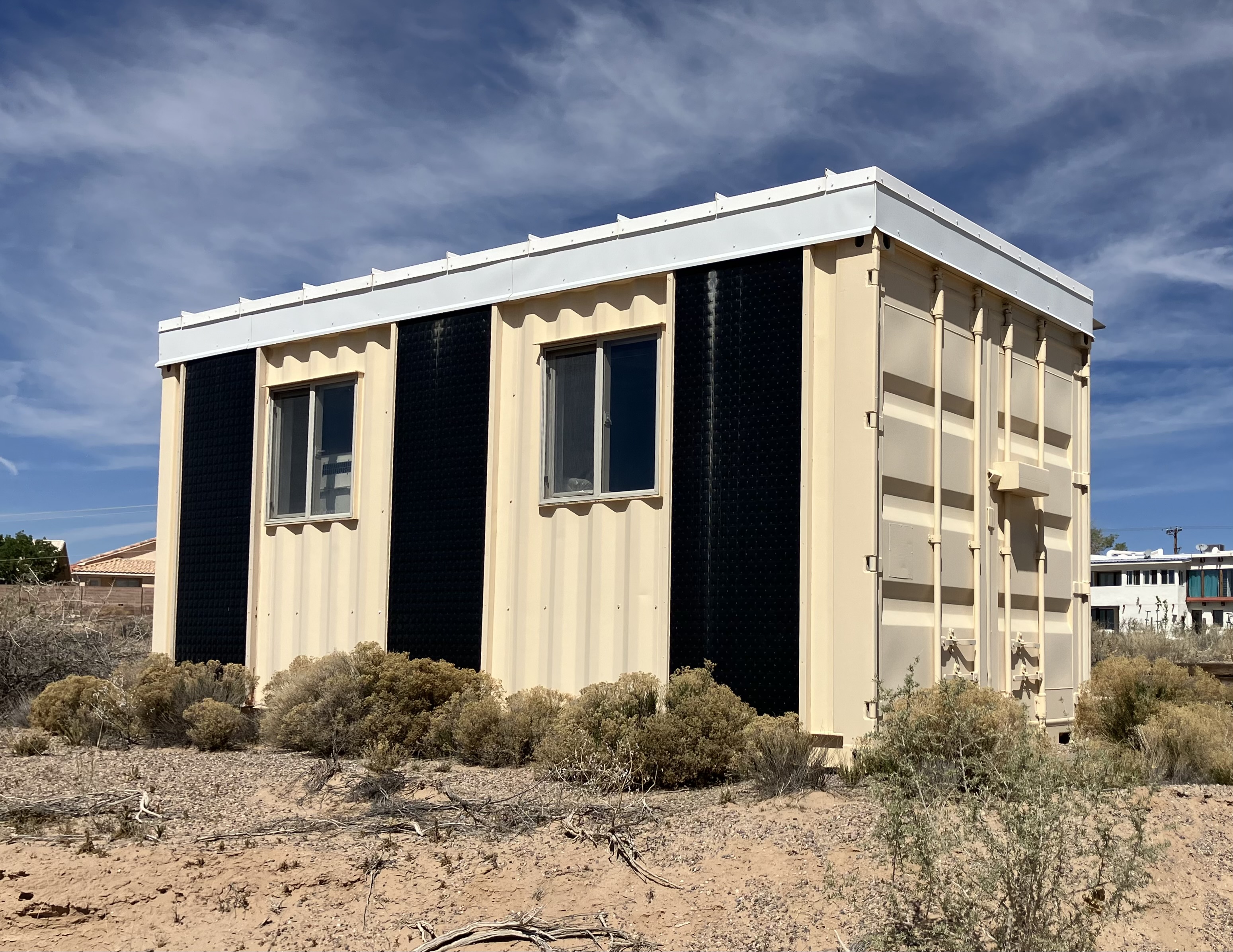 Exterior of a converted shipping container building in a New Mexico desert setting, with black thermal absorber panels covering the south-facing wall